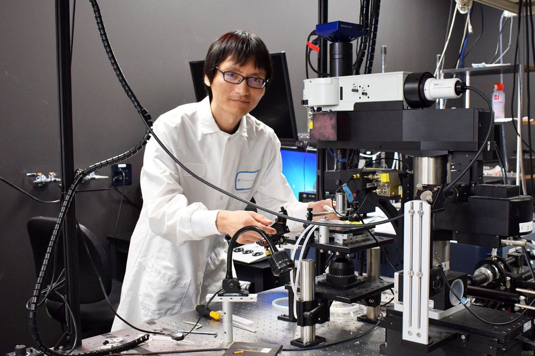 UF neuroscientist Ryoma Hattori in a labcoat looking at the camera over a complex laboratory microscope setup
