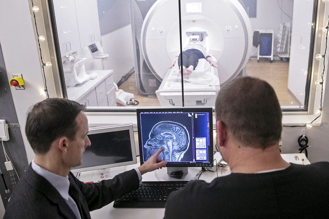 David Vaillancourt points at a screen showing an image of a brain MRI, with a patient in an MRI machine in the background