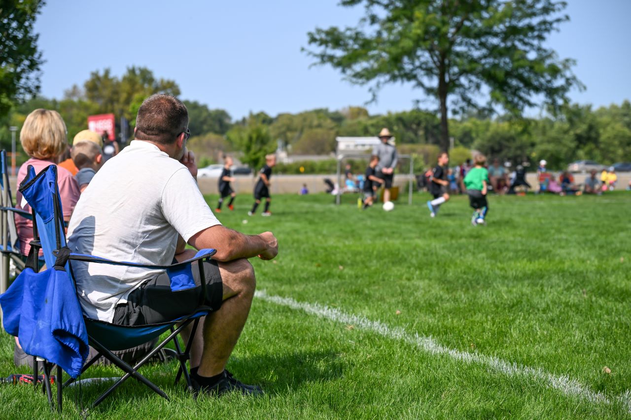 A father on a chair watches youth soccer outside