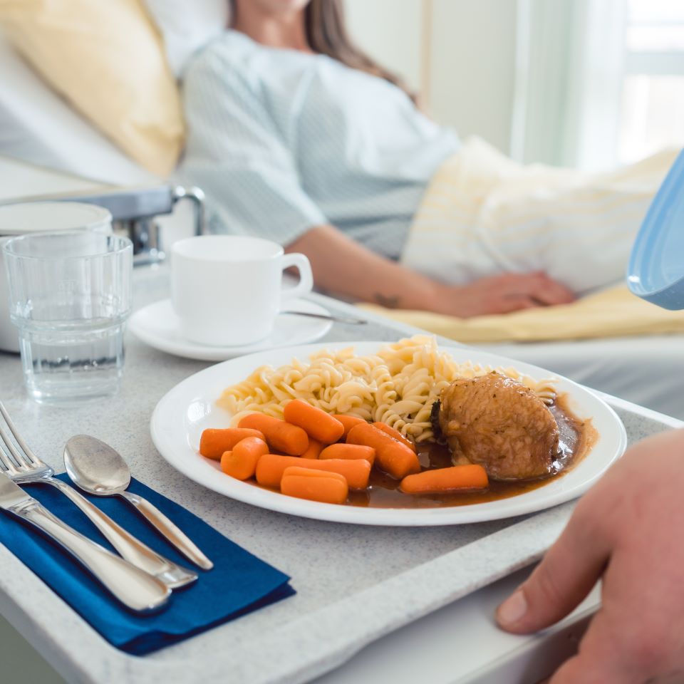 A tray of cafeteria-style food being delivered to a woman in a hospital bed