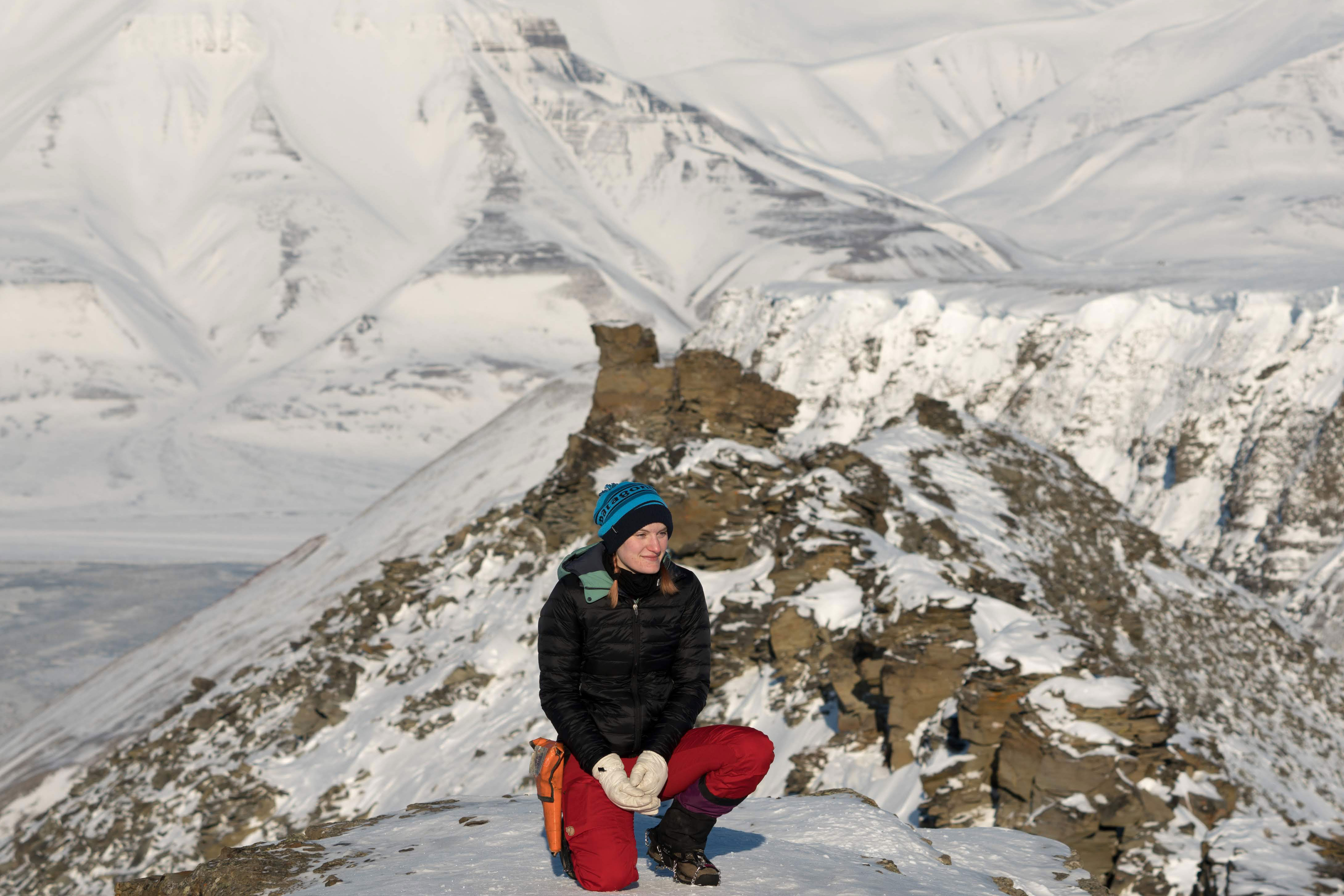 A woman crouches for a picture with an icy mountainous range in the background.