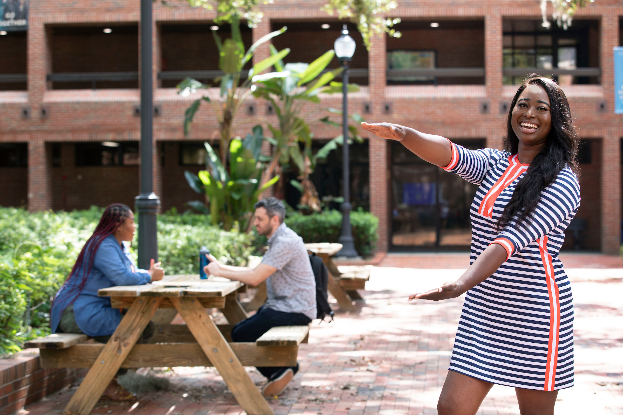A photo of a student doing the Gator chomp, with two students seated a picnic table in the background.