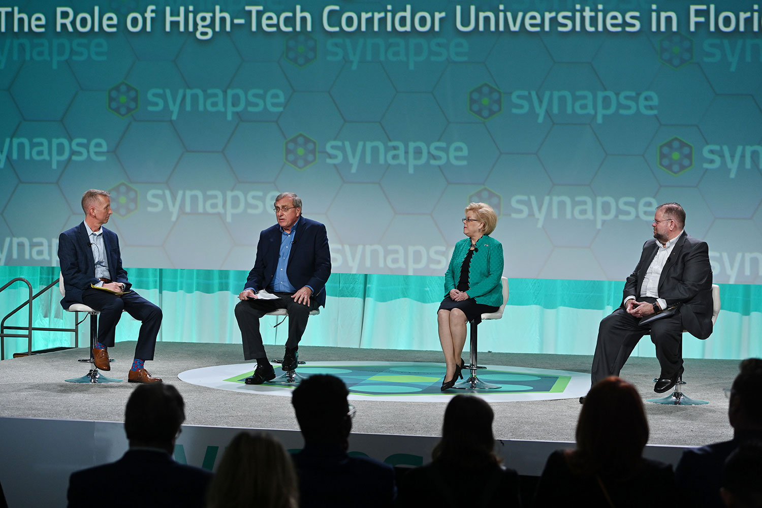 Four people sit on a stage engaging in a conversation while a crowd watches on