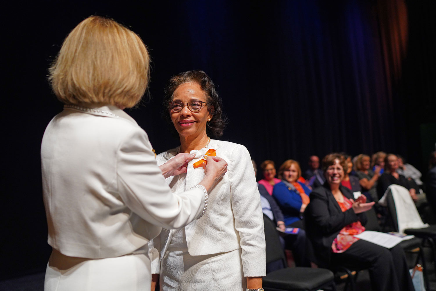 A woman is pinning another woman's shirt at a ceremony on stage