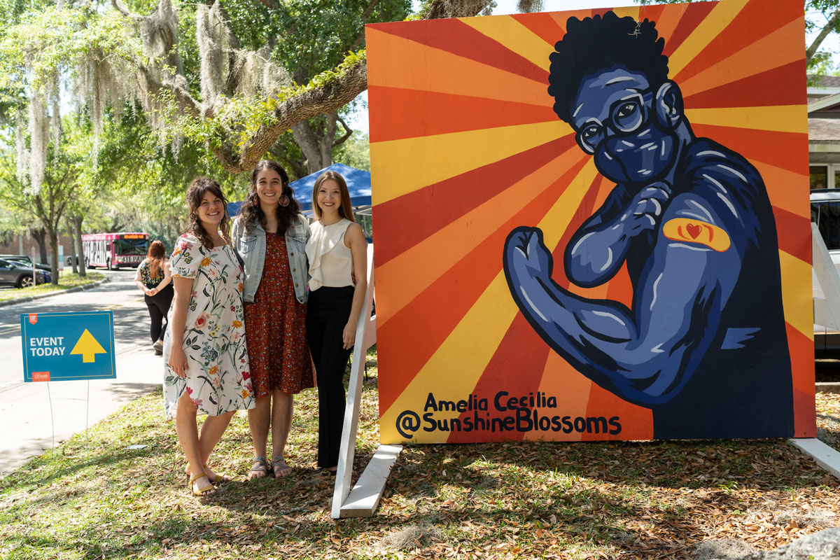 Three people pose next to a mural of a person flexing their bandaged arm
