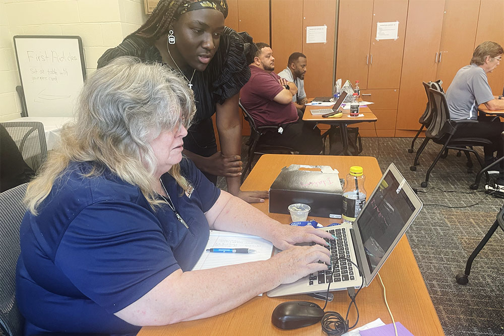 An engineering student peers over shoulder of high school teacher working on laptop.