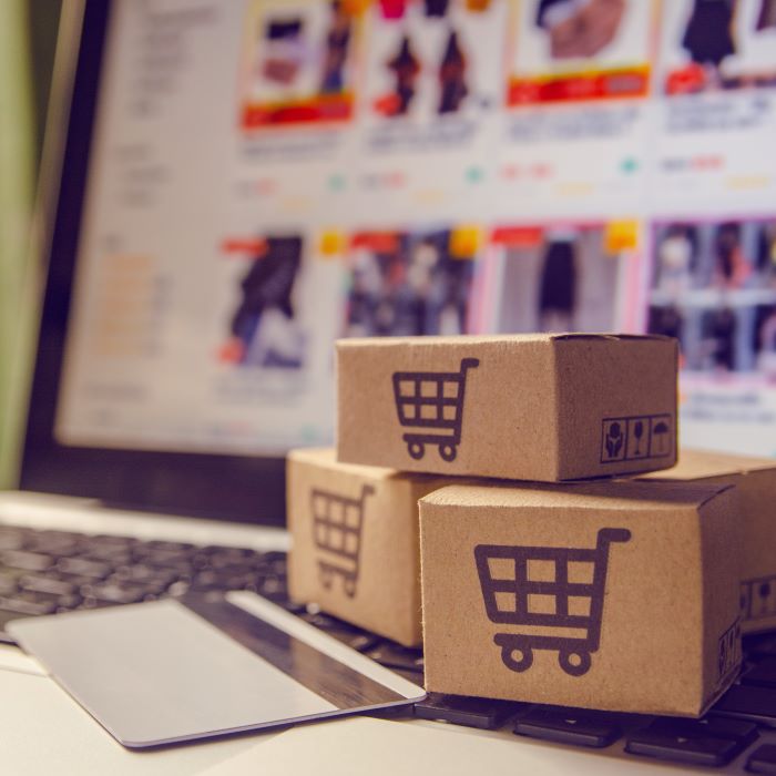 A laptop screen shows consumer products available for purchase. In the foreground, miniature shipping boxes are stacked on top of the laptop near a credit card.