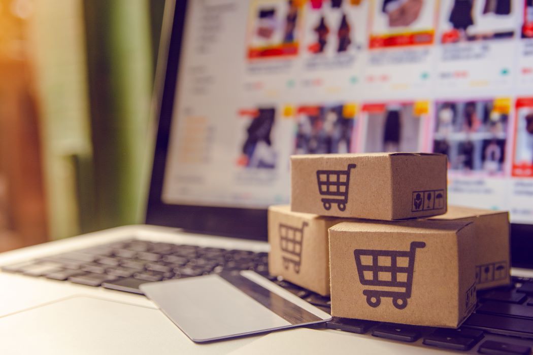 A laptop screen shows consumer products available for purchase. In the foreground, miniature shipping boxes are stacked on top of the laptop near a credit card.