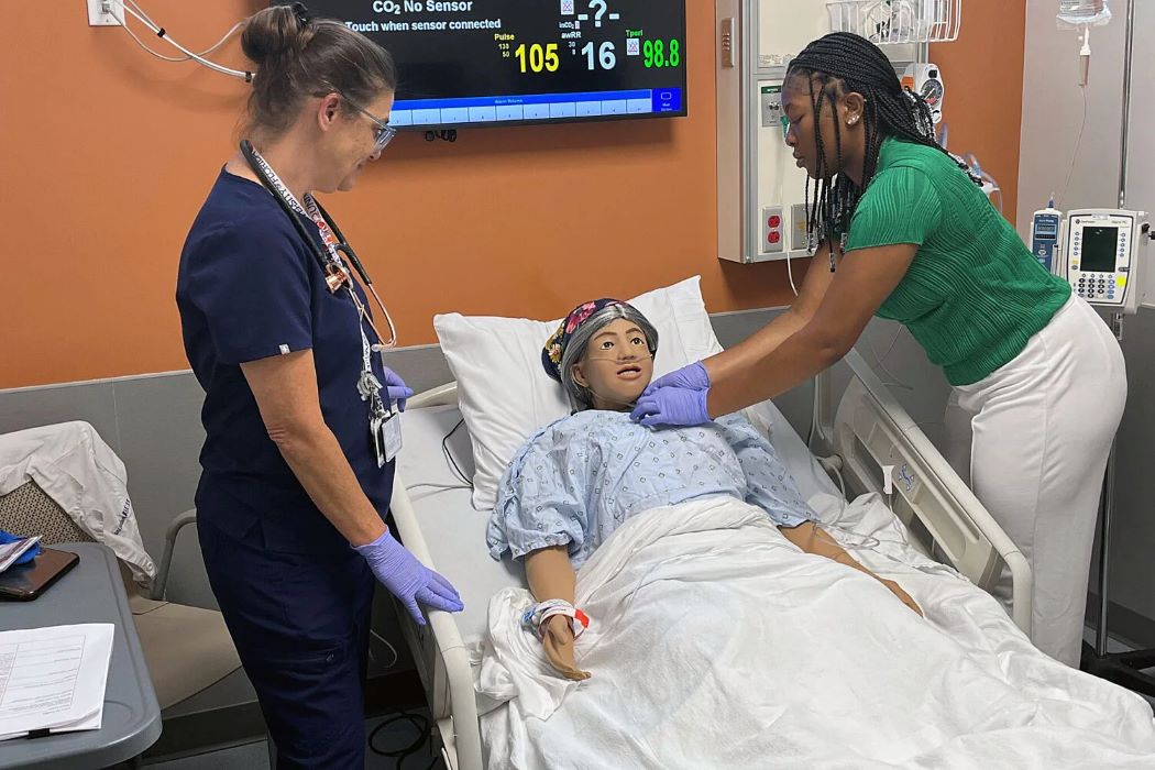 A student tends to a dummy patient in a hospital bed as a healthcare trainer looks on
