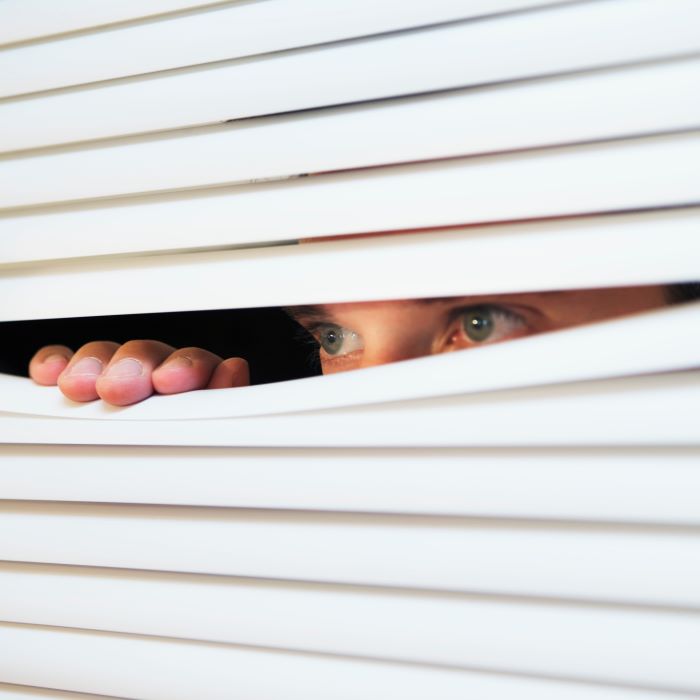 A man's eyes peek from behind venetian blinds as his hand pulls the blinds apart