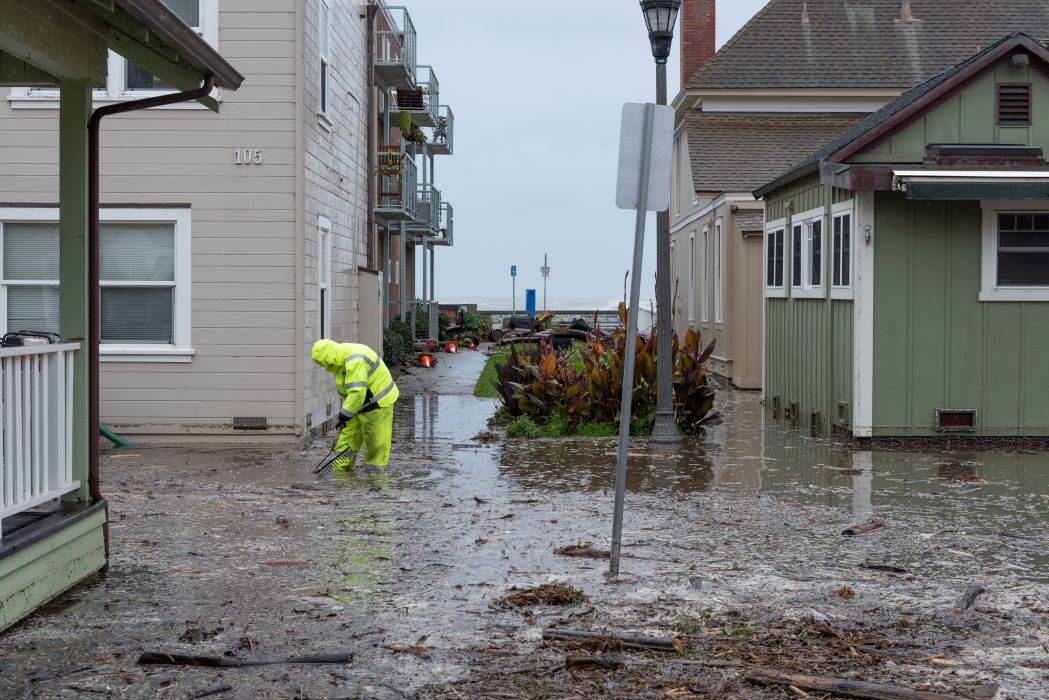 A person in neon visibility gear holds a rake and peers into the flood waters between multiple apartment buildings