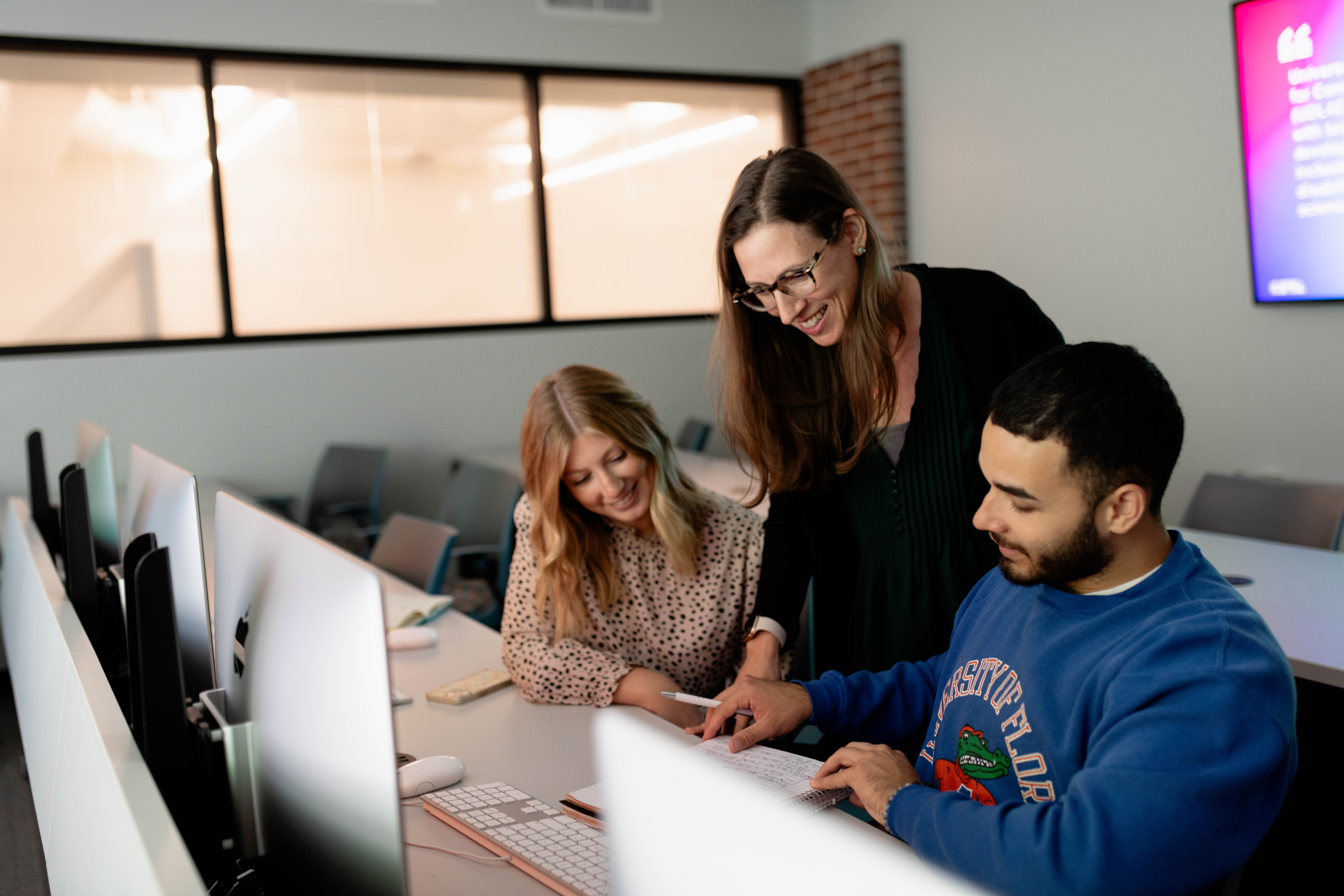 A female instructor stands behind two students sitting at computers.