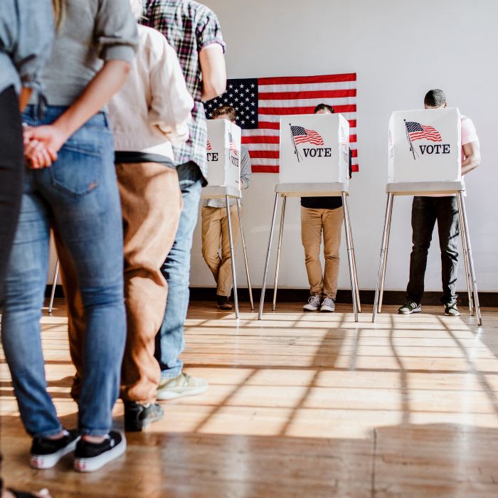 A voting center with an American flag hanging on the wall in the background, three voters obscured behind privacy screens and a line of people waiting to vote in the foreground
