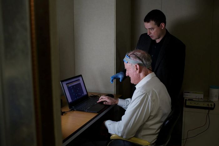 An older adult sits at a computer wearing an electrode cap while a main standing behind him points with a gloved hand at the computer screen