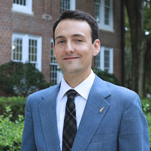 A headshot of male faculty member dressed in a suit and tie standing in front of a building.