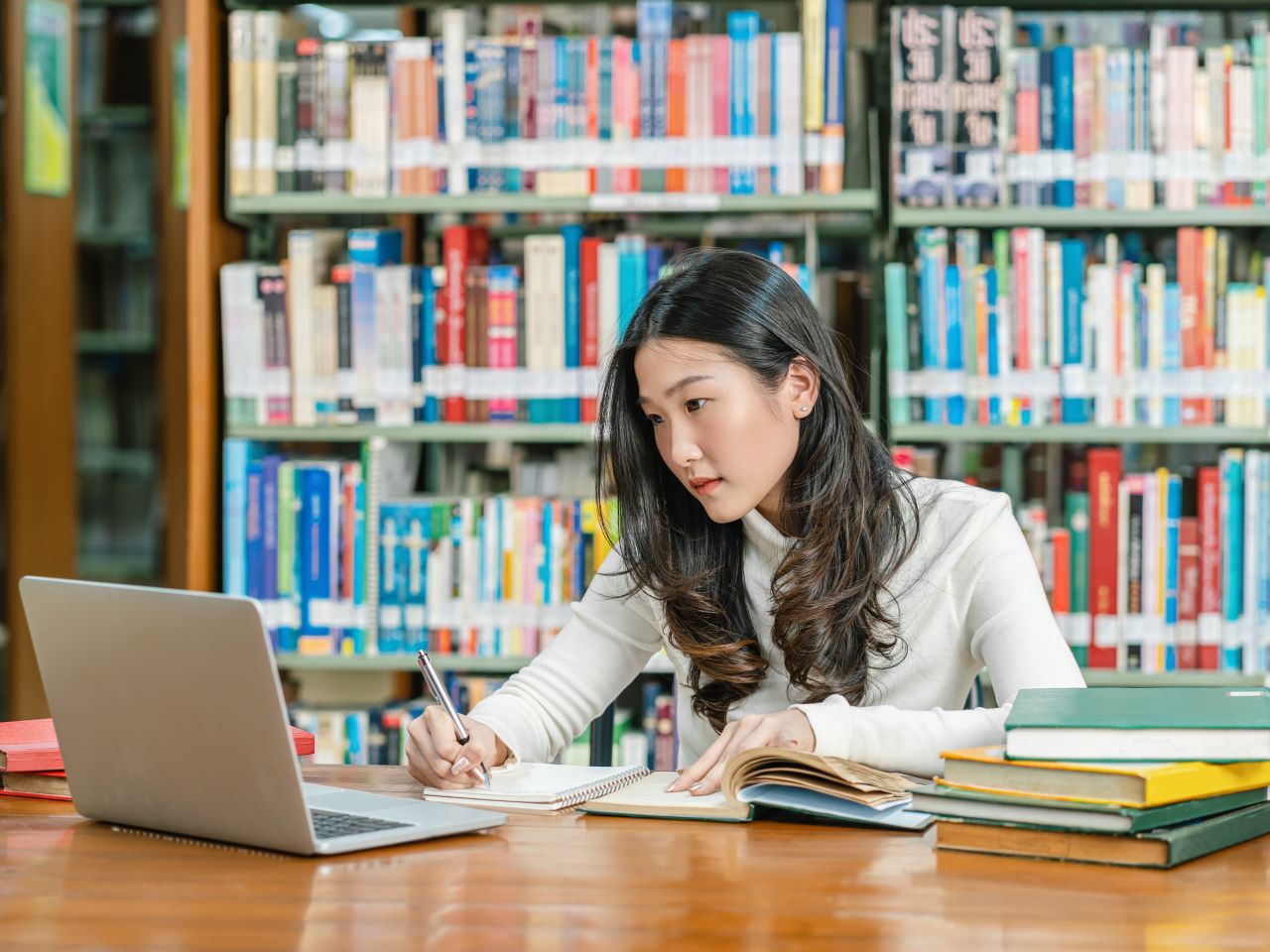 A student sitting at a library table with a stack of books and a laptop