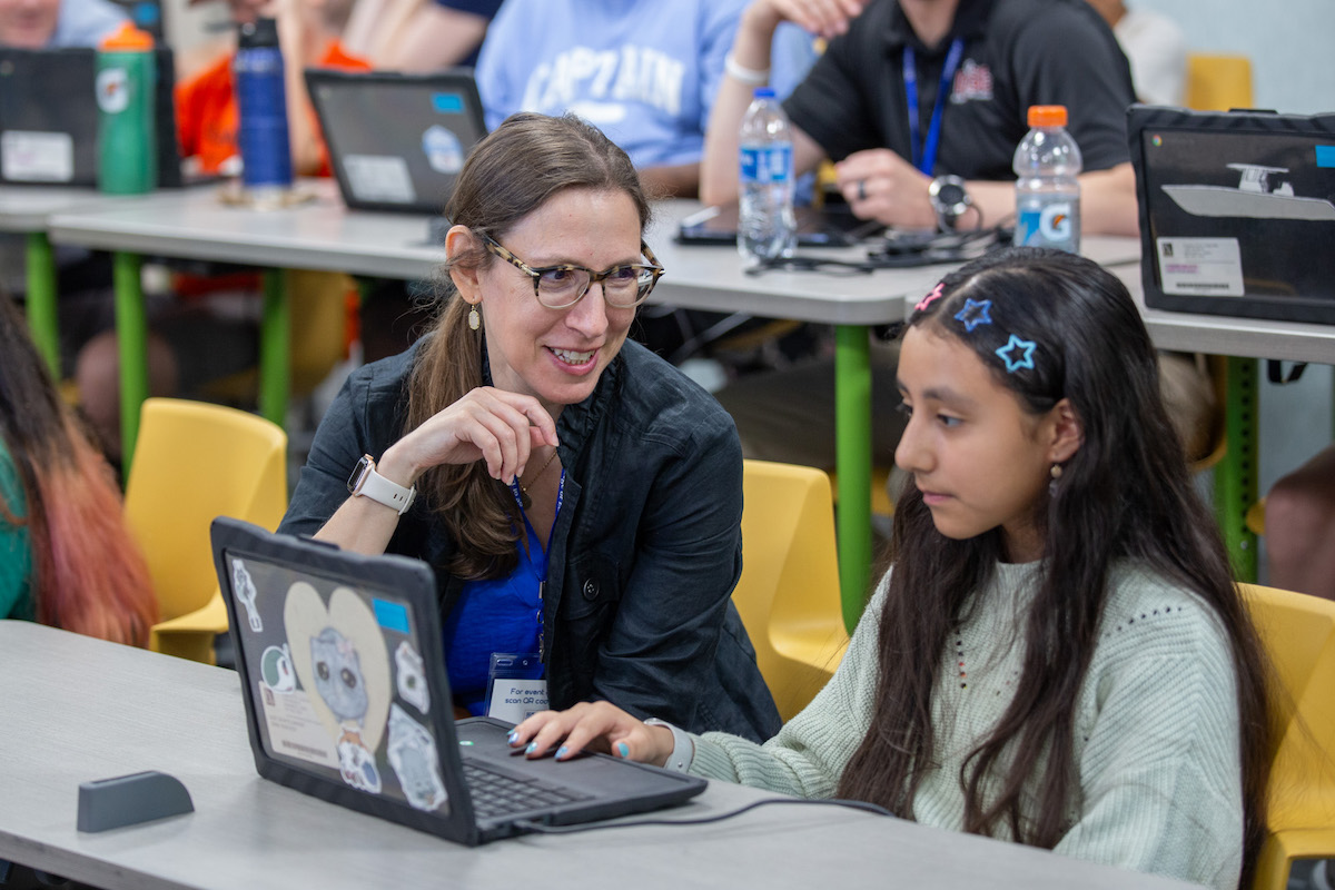 A female teacher sitting next to a student on a laptop helping her.