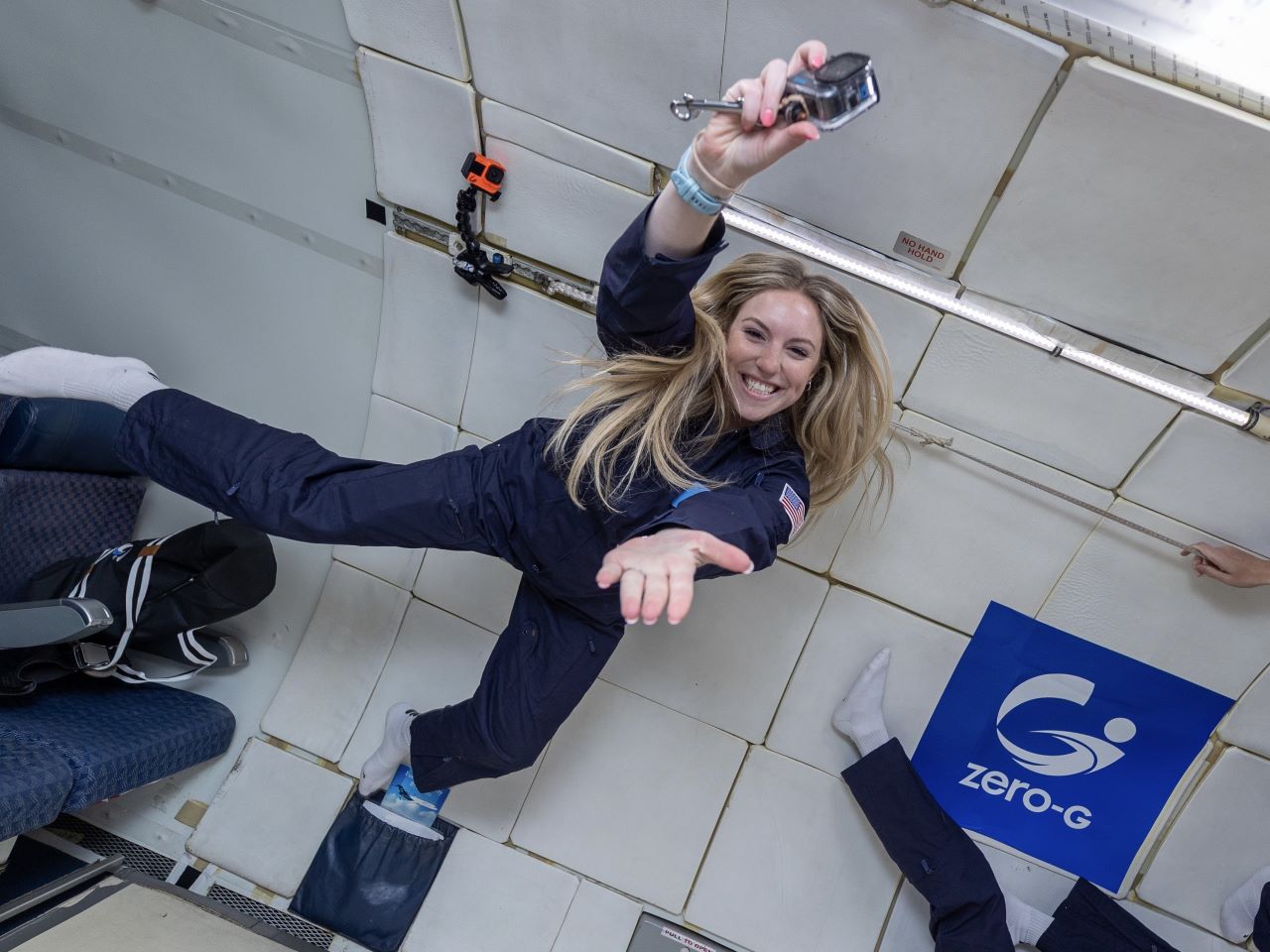 Hannah Roberts wears a blue flight suit and does the Gator Chomp while floating inside a parabolic flight plane