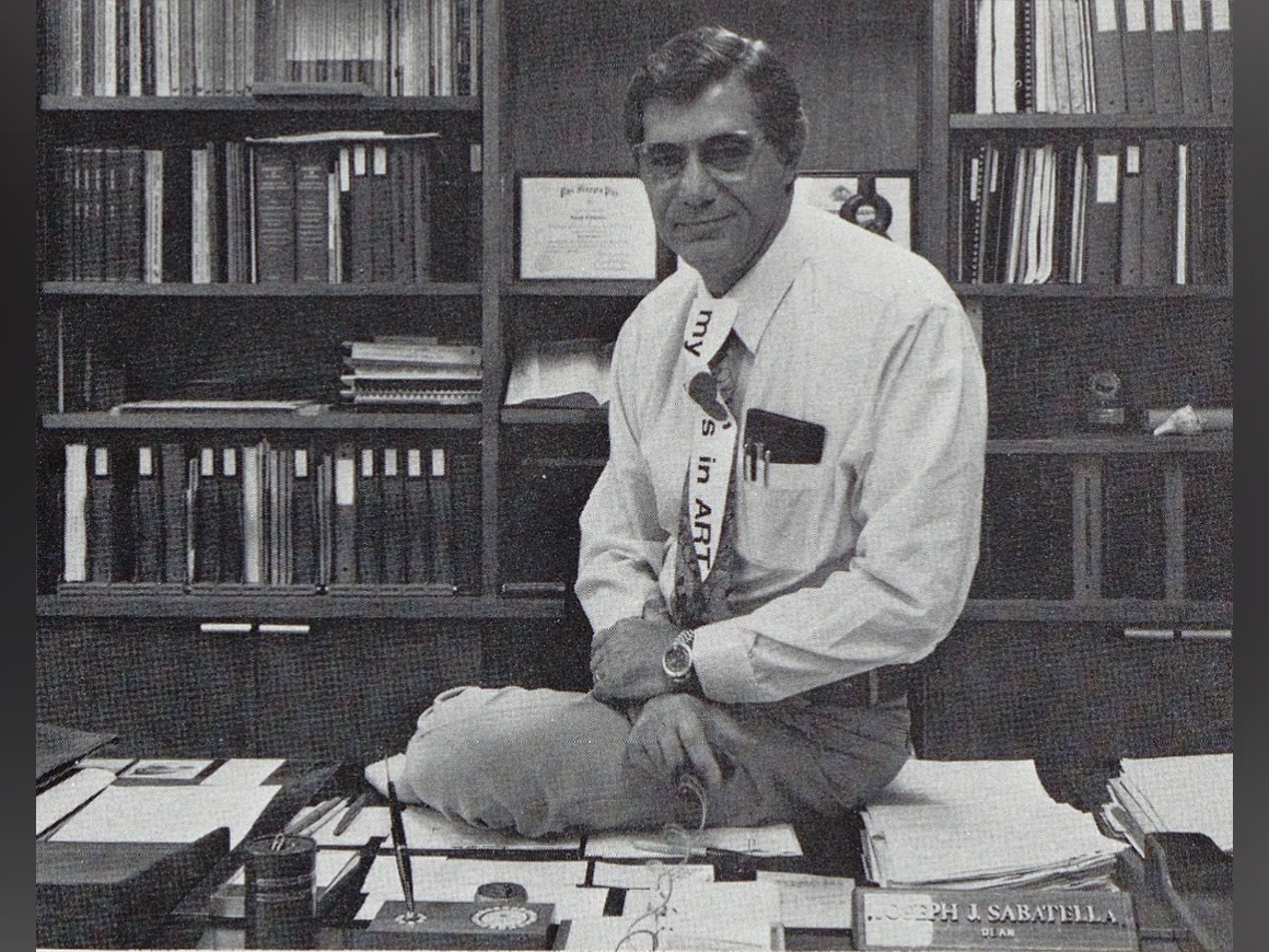 A black and white photograph of Dean Joseph Sabatella sitting on a desk